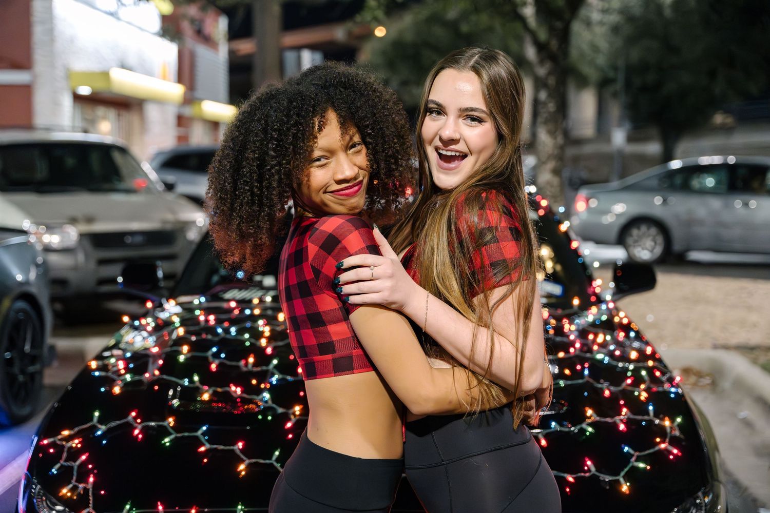 Two friends in matching buffalo plaid tops pose together with holiday lights in background.