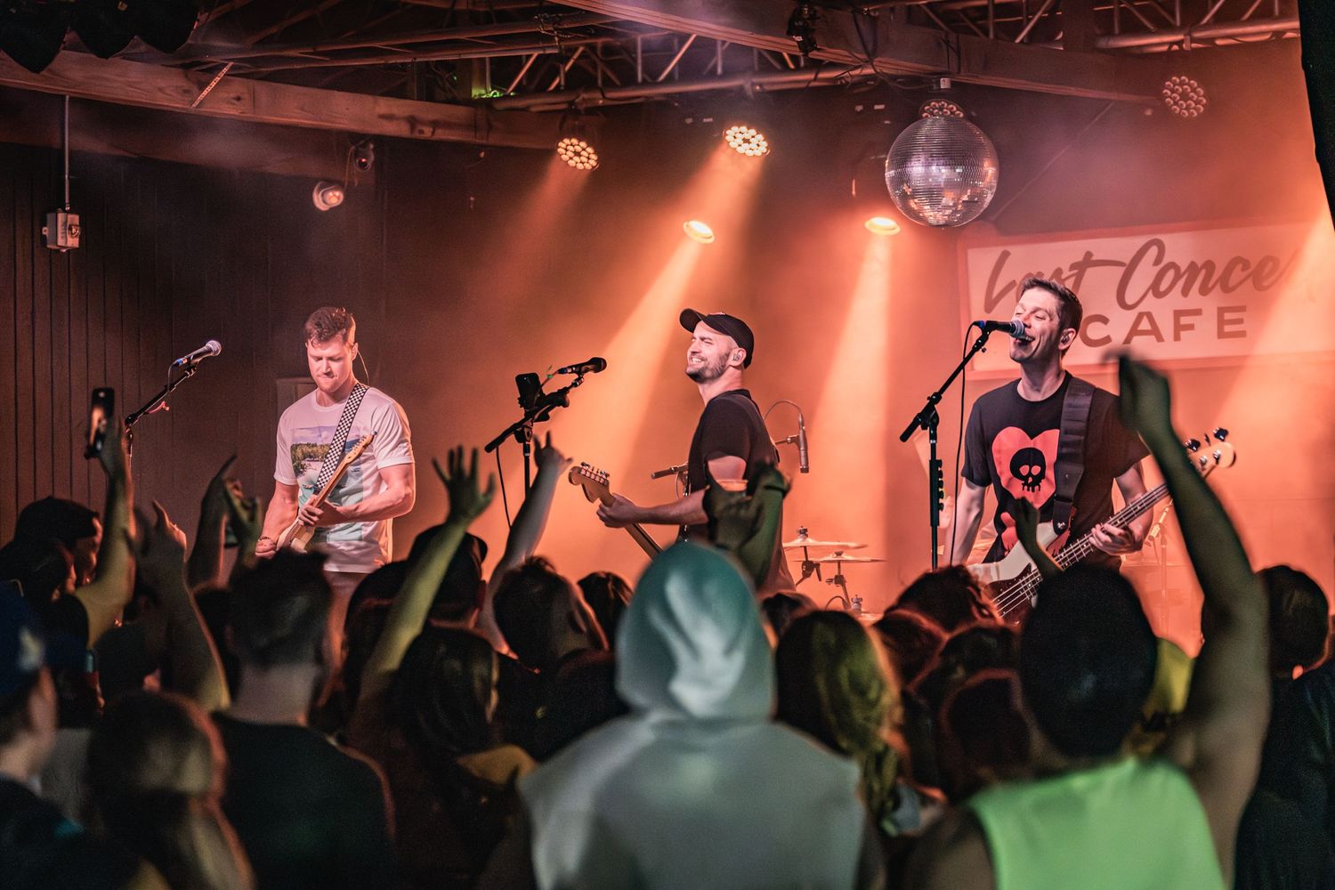 Band performing on stage with spotlights and disco ball while crowd cheers at concert venue.