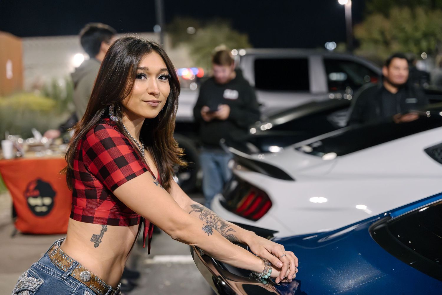 Woman in plaid crop top posing with blue car at night meet.