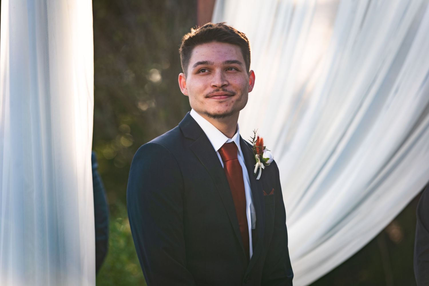 Wedding party member poses in navy suit with red tie against white drapery backdrop.
