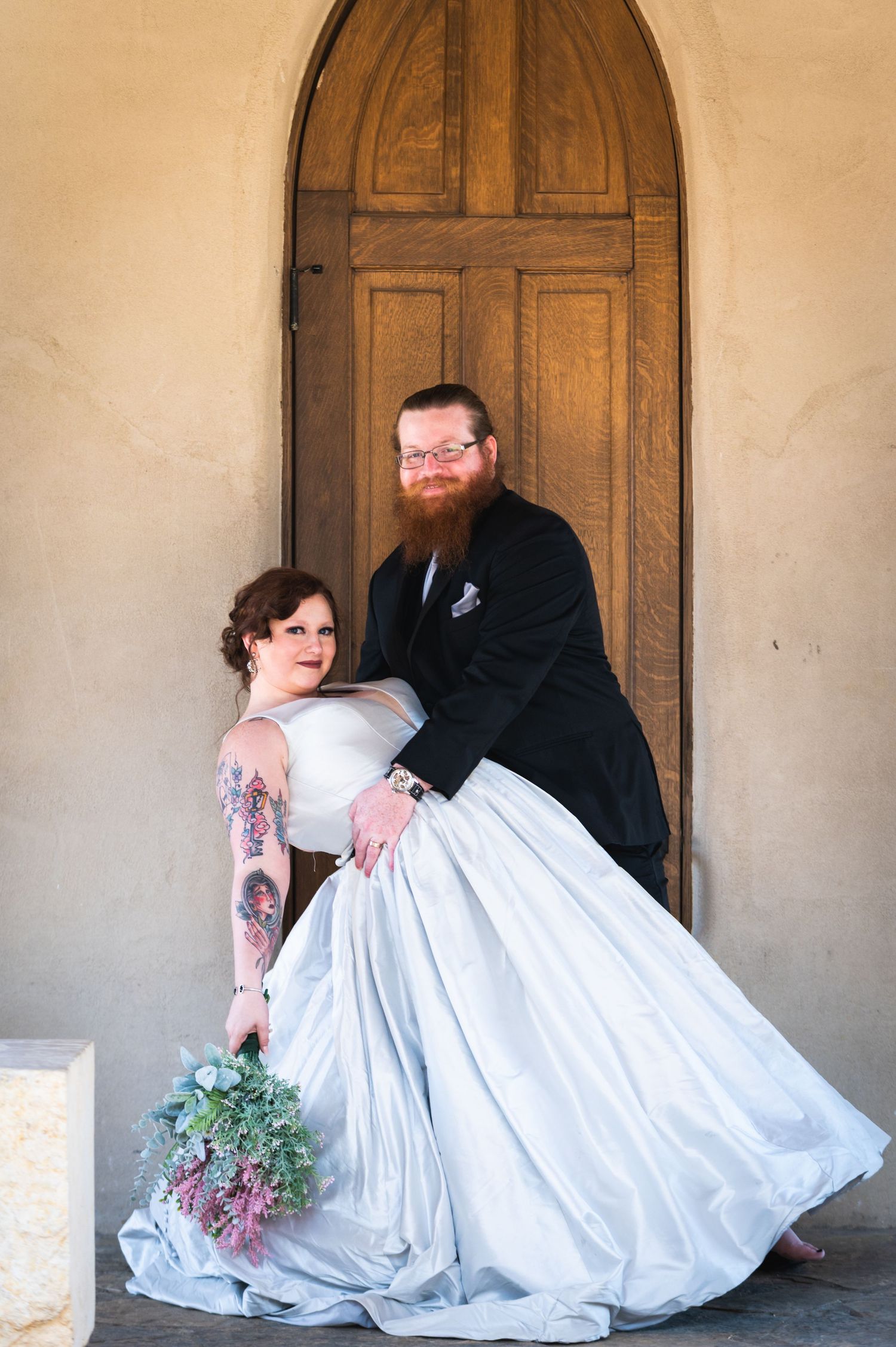 Wedding couple poses by wooden church door with bride holding blue and purple bouquet.