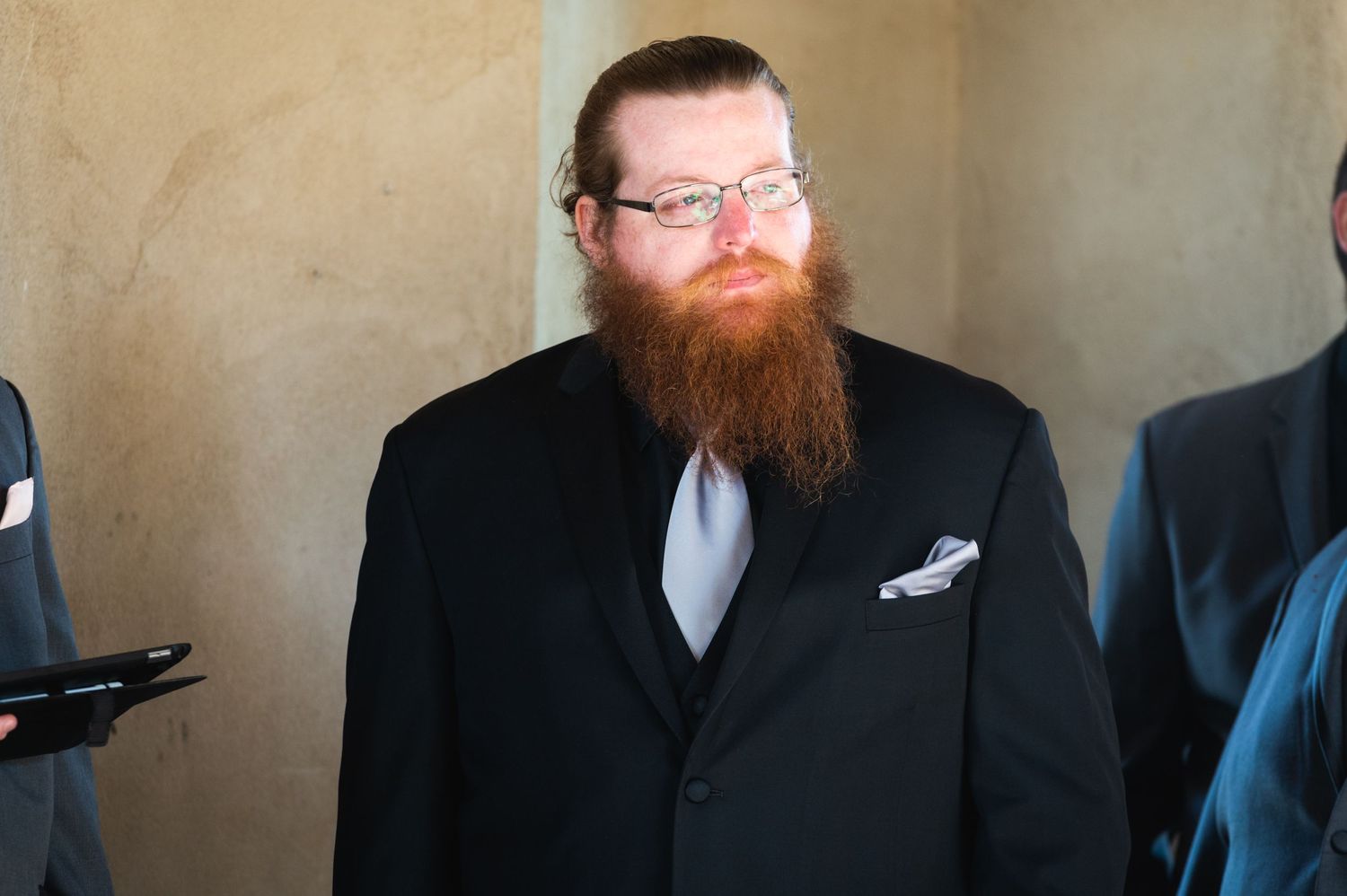 A person in formal attire with a red beard and glasses stands during a wedding ceremony.