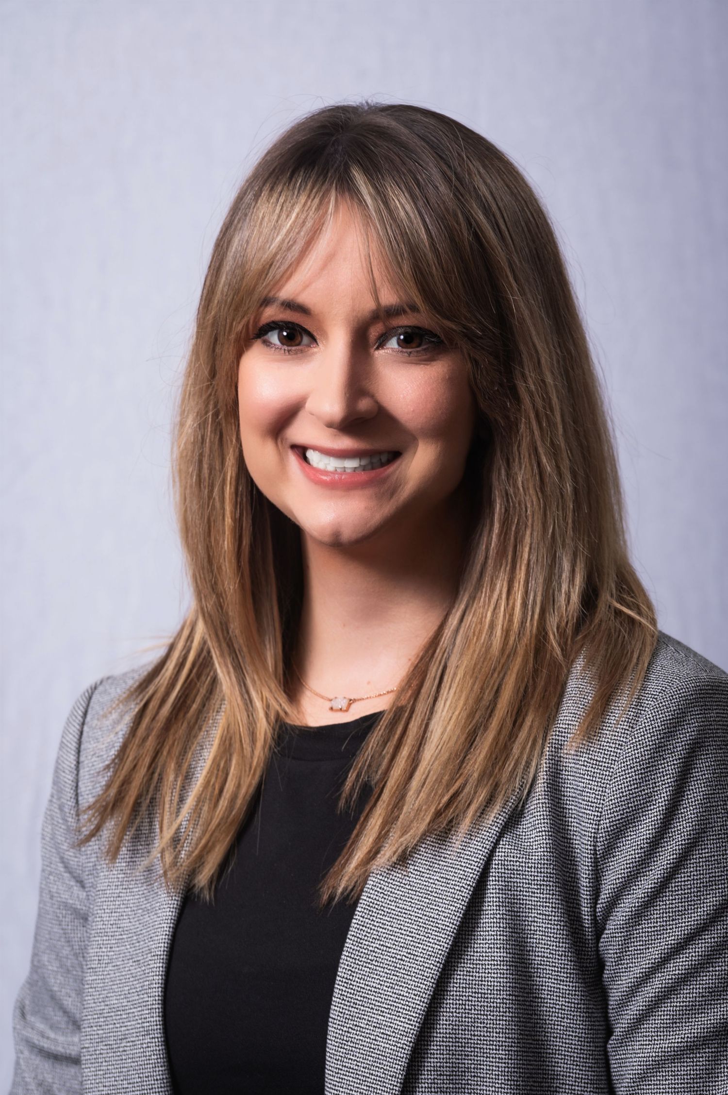 Professional headshot portrait of someone in business attire with long hair and bangs against a gray backdrop.