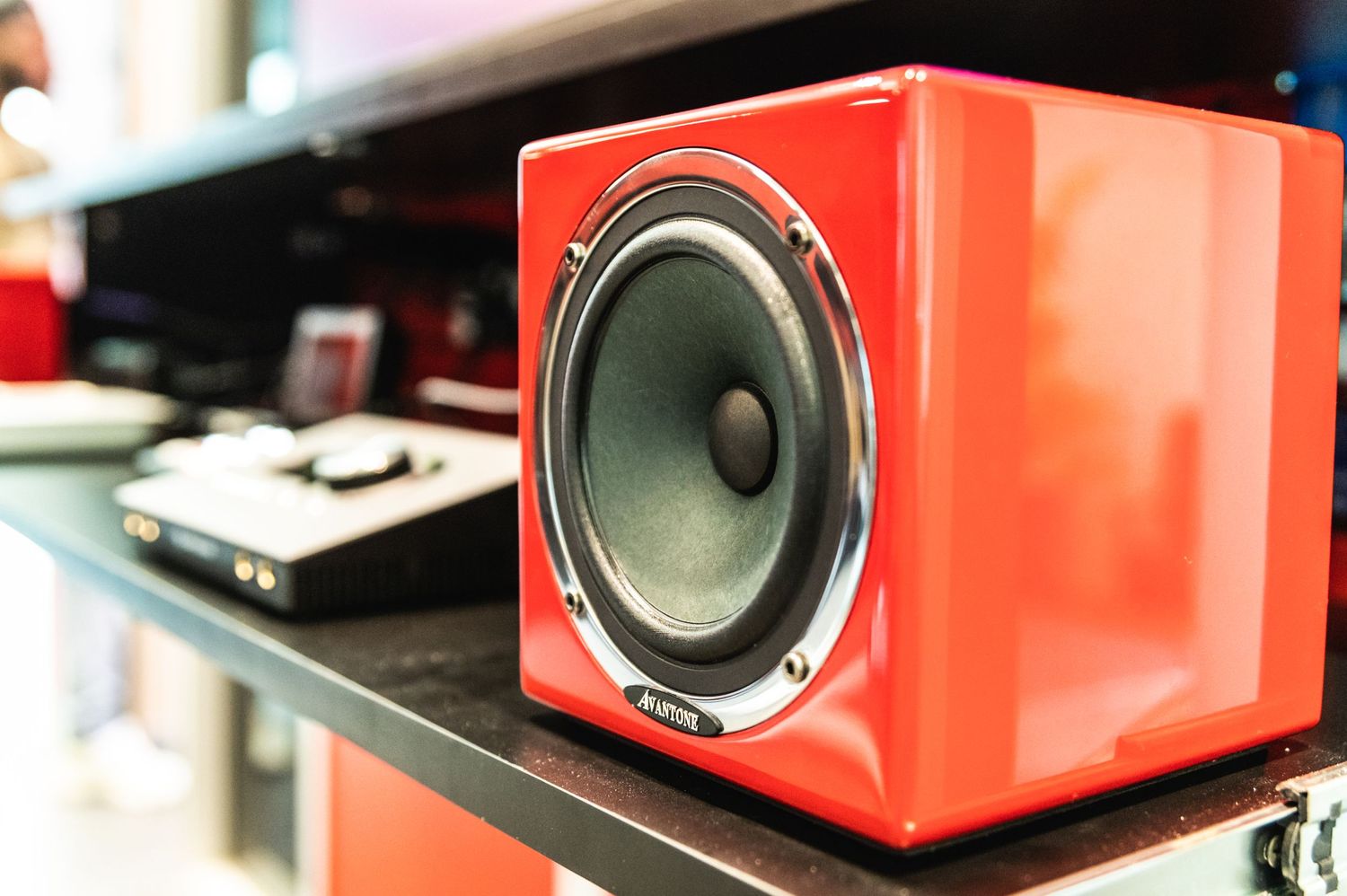 Red cube speaker with visible woofer sits on black shelf in audio setup.