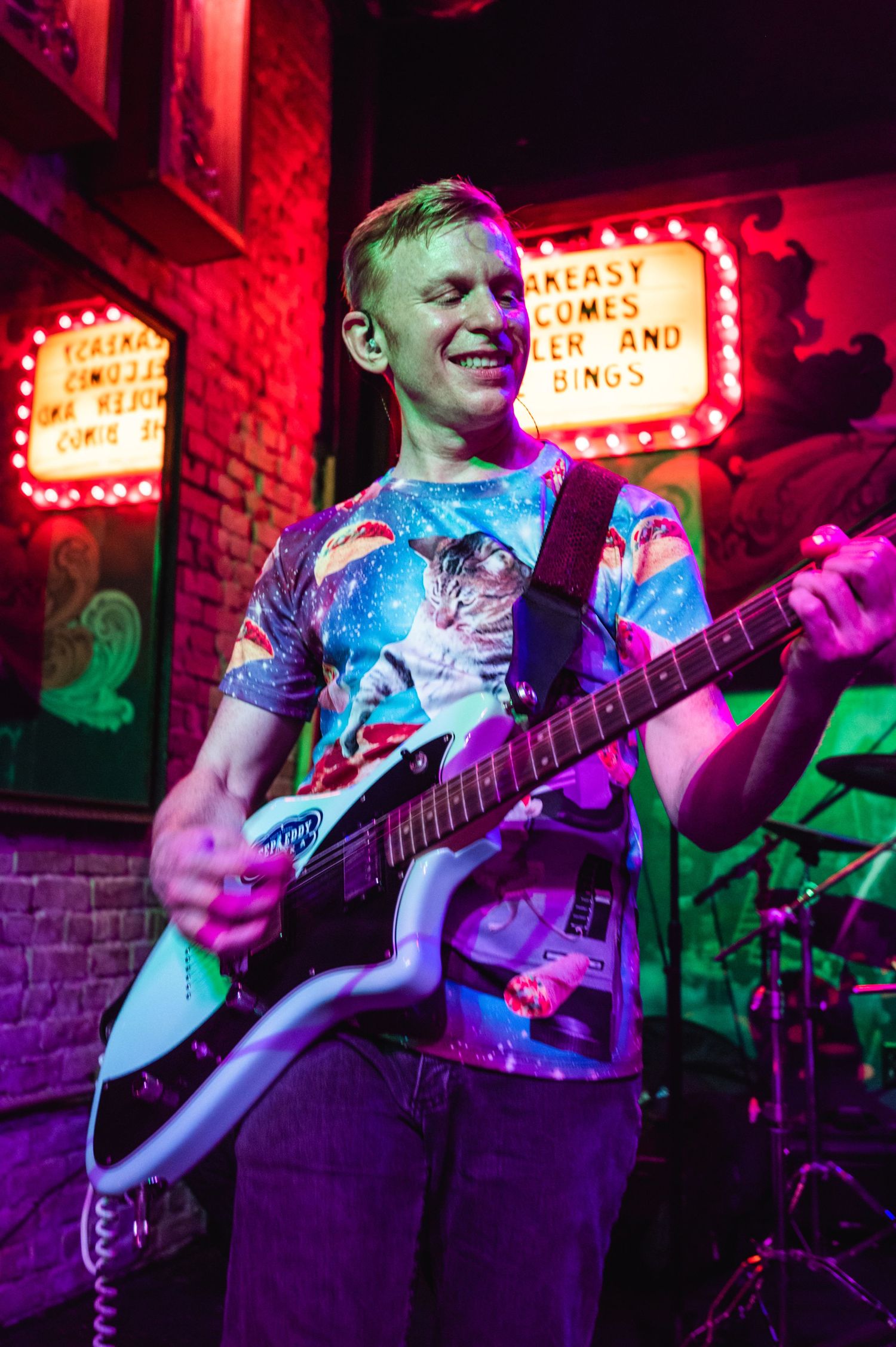 Guitarist wearing vibrant tie-dye shirt performs under neon lights at a small club.
