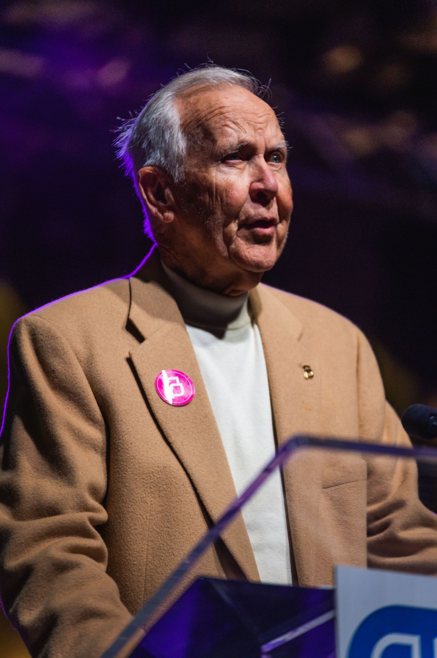 Speaker wearing tan blazer and white turtleneck addresses crowd from illuminated podium.