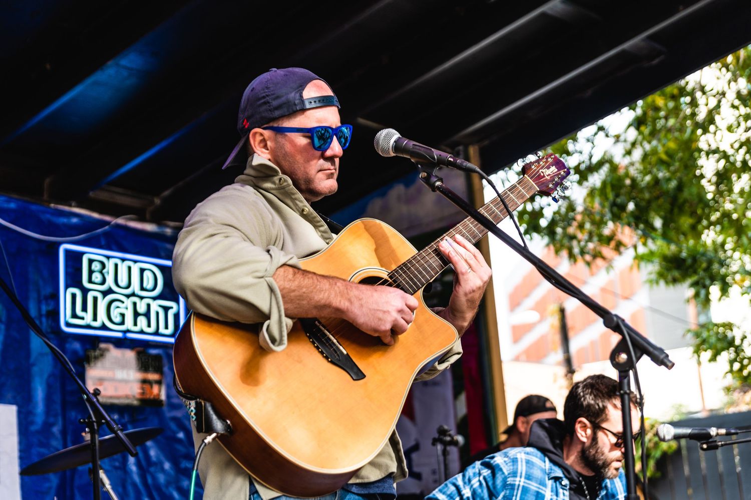 Musician performs with acoustic guitar on outdoor stage with Bud Light banner in background.