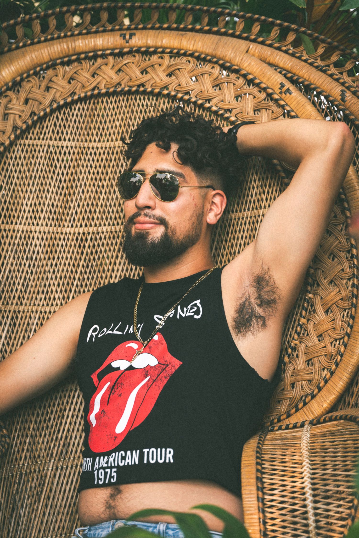 Person in Rolling Stones tank top relaxes in wicker peacock chair wearing sunglasses.