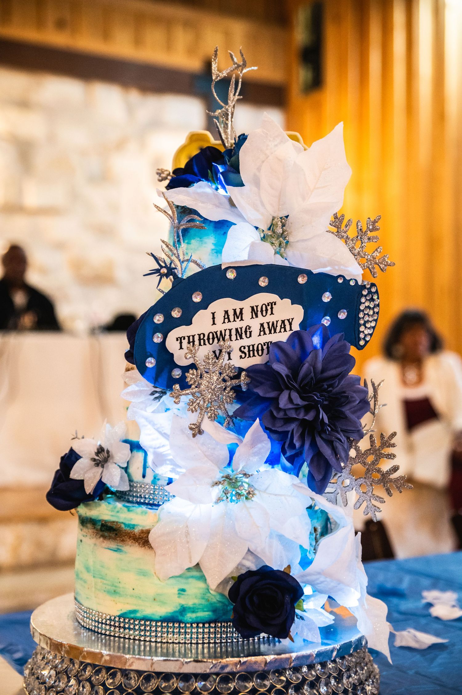 Whimsical wedding cake with blue and white flowers featuring Hamilton musical theme and decorative text.