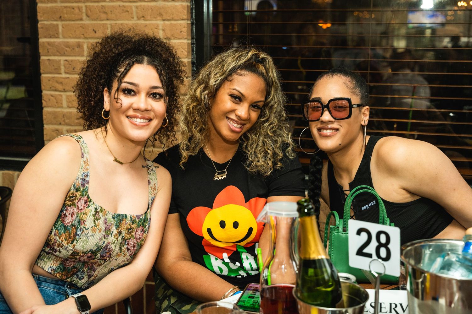 Three friends enjoying drinks and smiles at a casual restaurant gathering with a colorful emoji toy on the table.