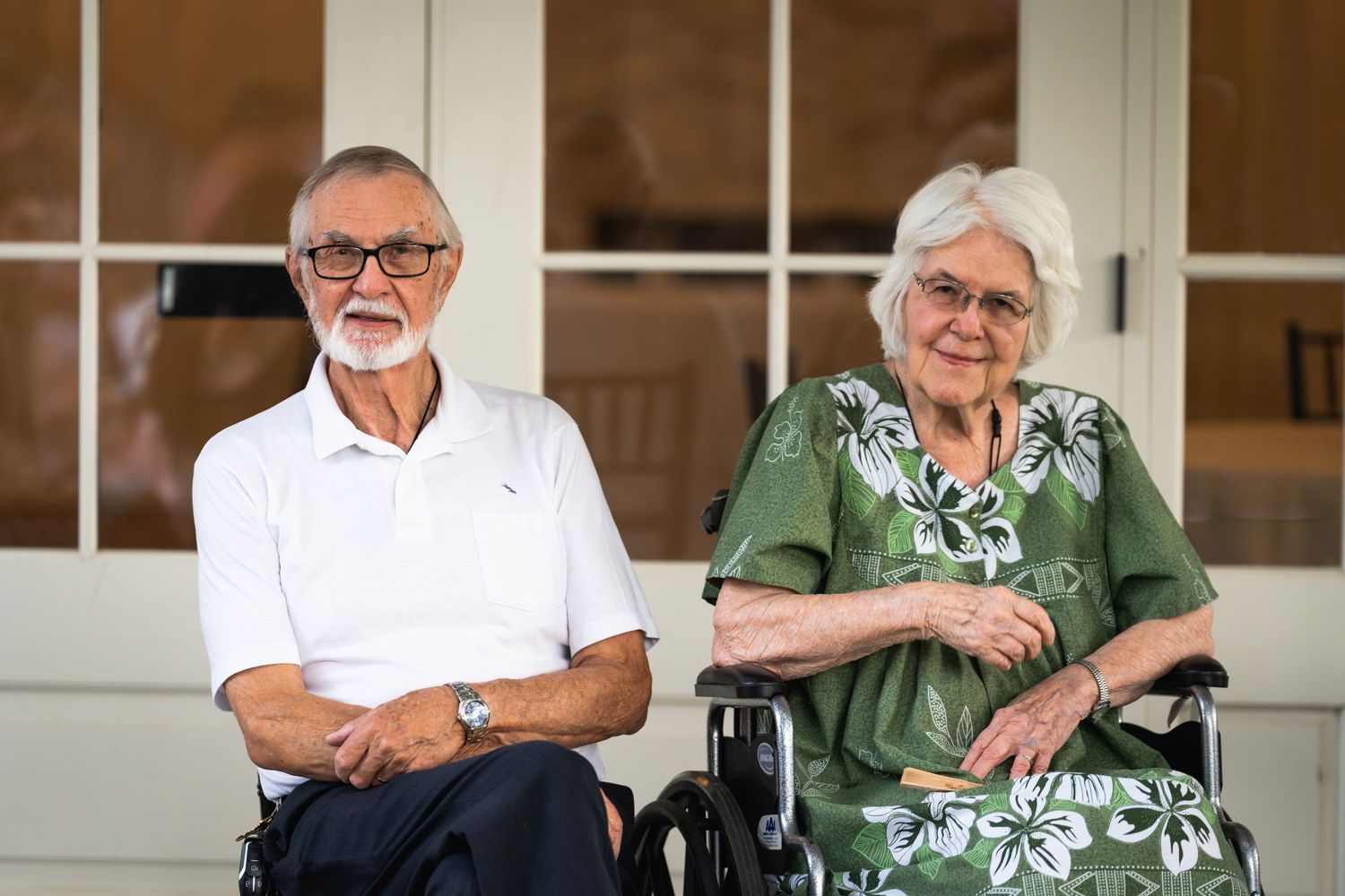 Elderly couple sitting on porch, woman in wheelchair wearing green floral dress next to man in white polo shirt.
