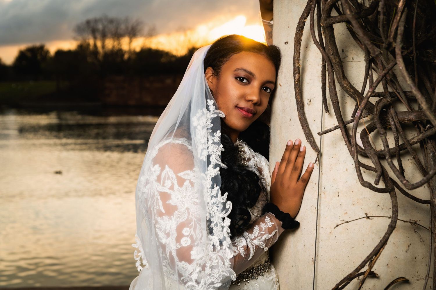 Bride in delicate lace gown poses against wall during golden hour by lake.