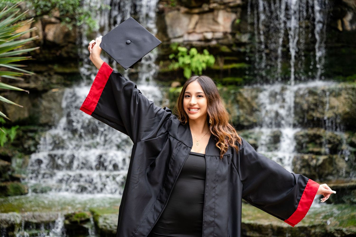 Joyful graduate in black gown celebrating with arms raised by waterfall.
