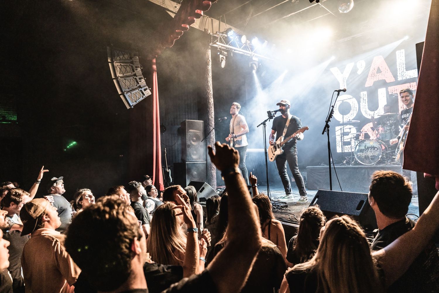 Enthusiastic concert crowd cheering at live rock show with band performing on stage under bright lights.