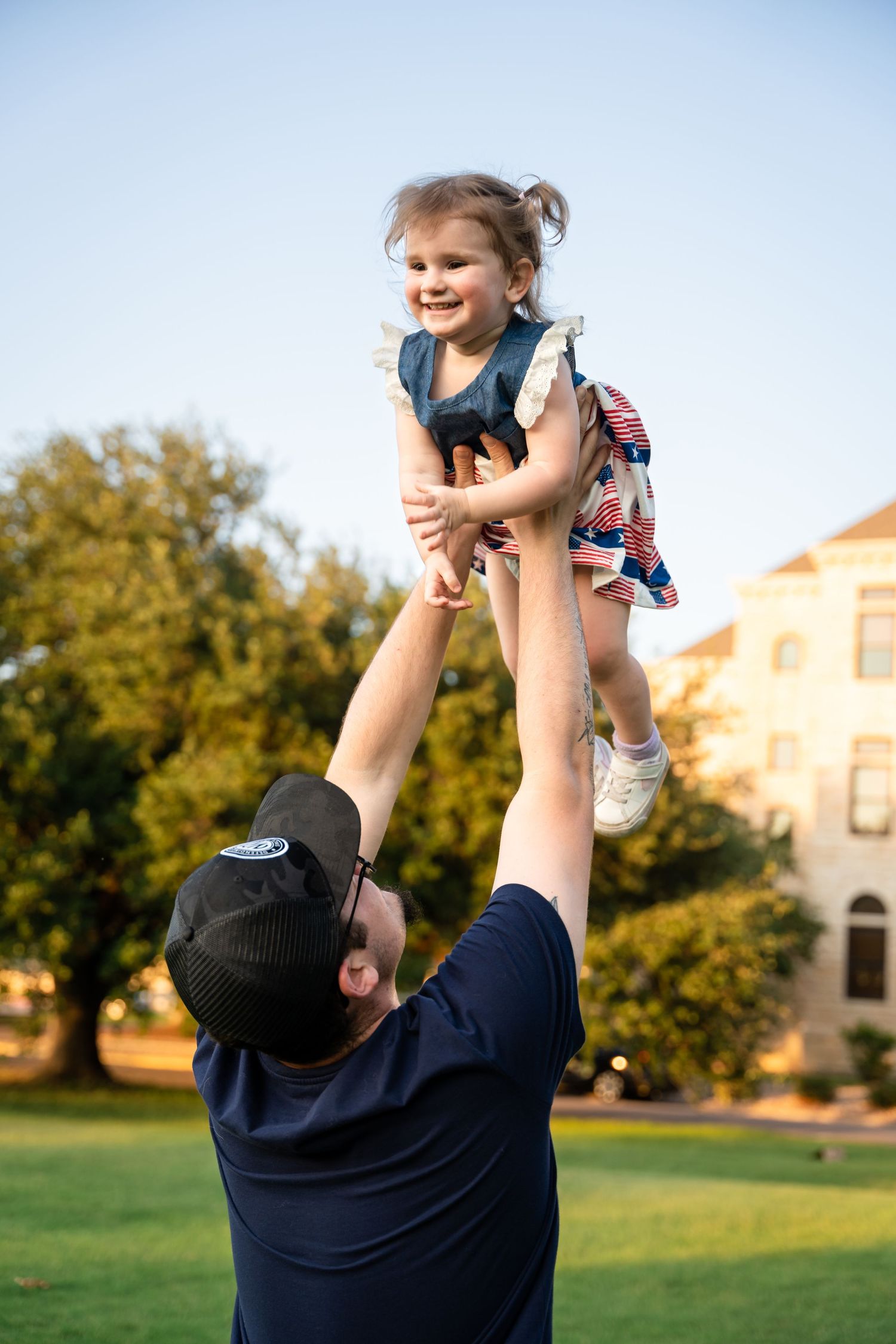 A playful lifting moment captured against a blue sky with trees and buildings in the background.
