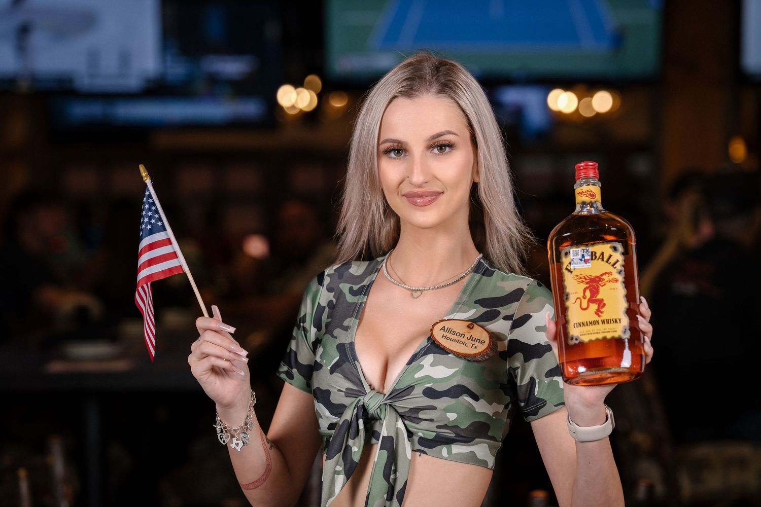 Model in camo print top holds American flag and whiskey bottle in restaurant setting.