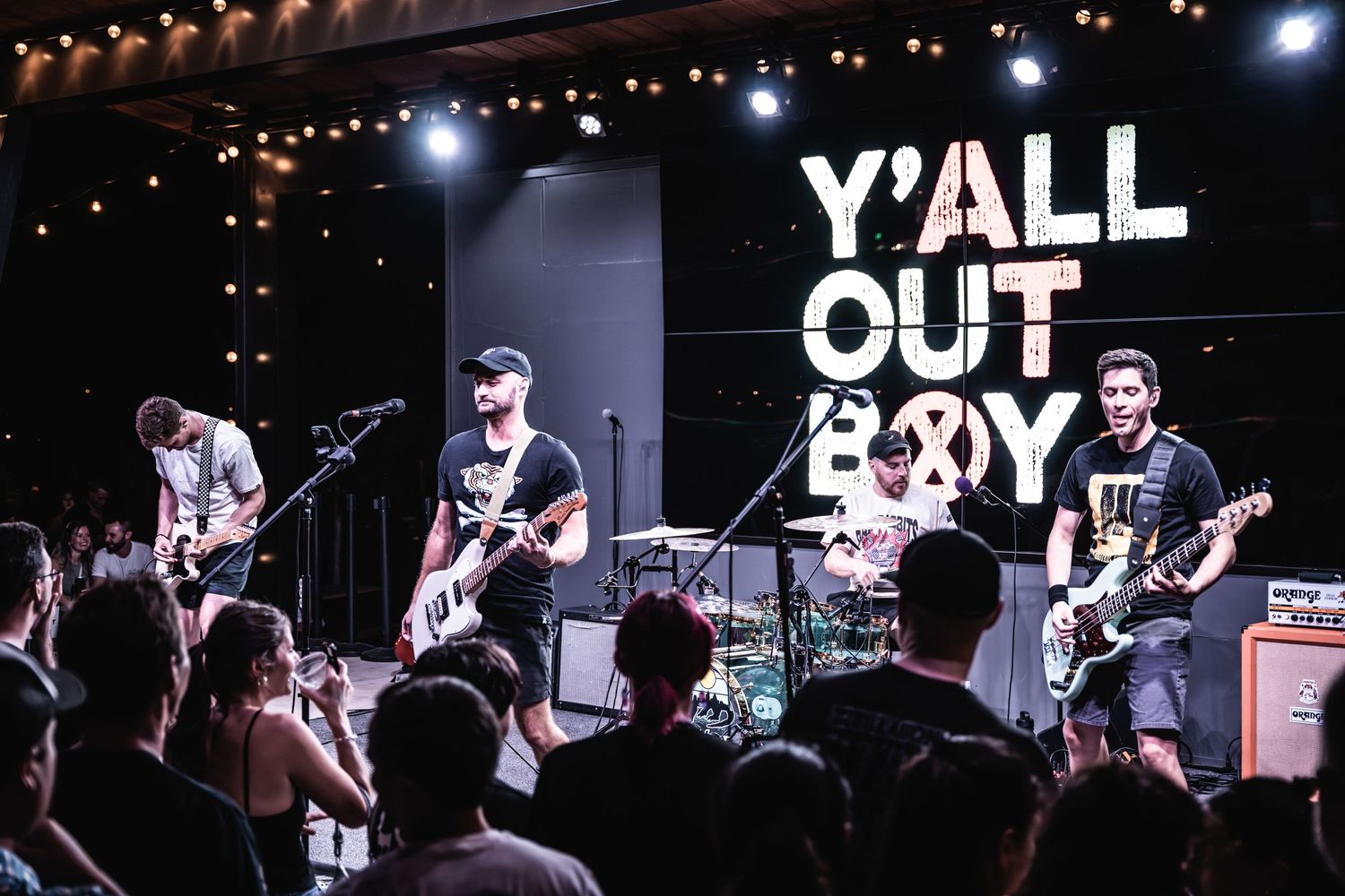 A band performs on stage under the Y'all Out Boy sign with string lights overhead.