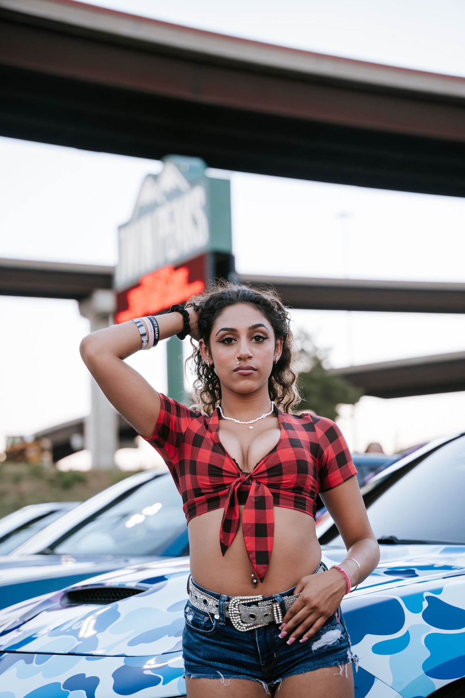 Fashion photo shoot with cars and plaid crop top styling against urban backdrop.