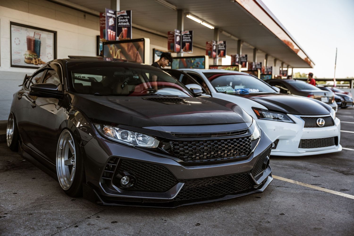 Modified black Honda Civic and white Lexus cars parked at a shopping center with custom wheels and lowered suspension.
