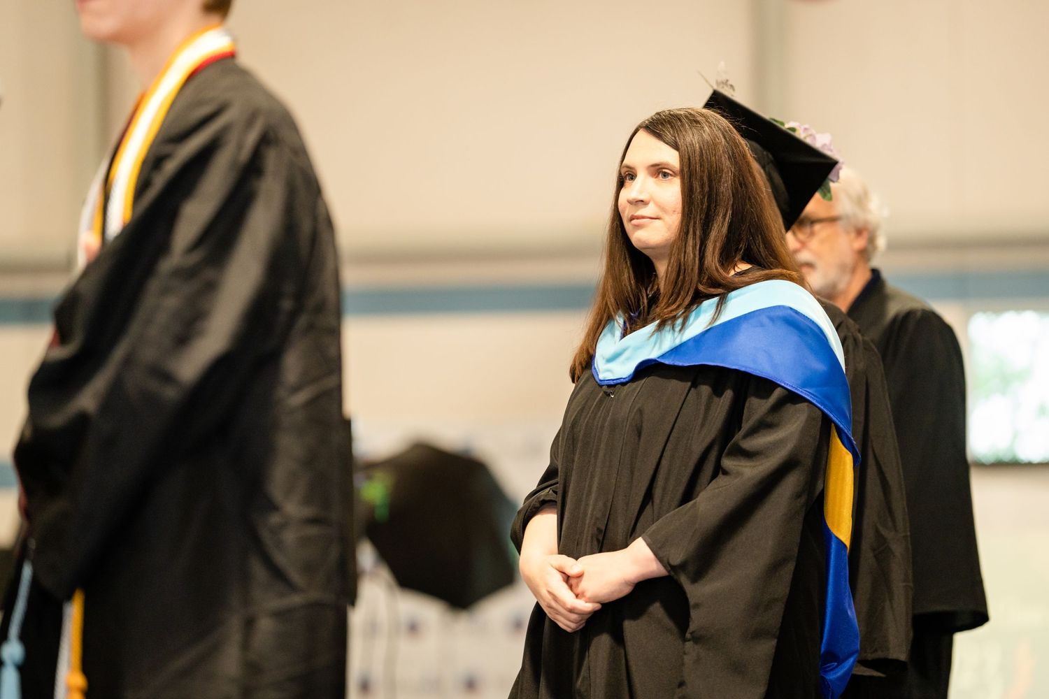 Graduate in traditional black cap and gown with blue honors stole stands during ceremony.