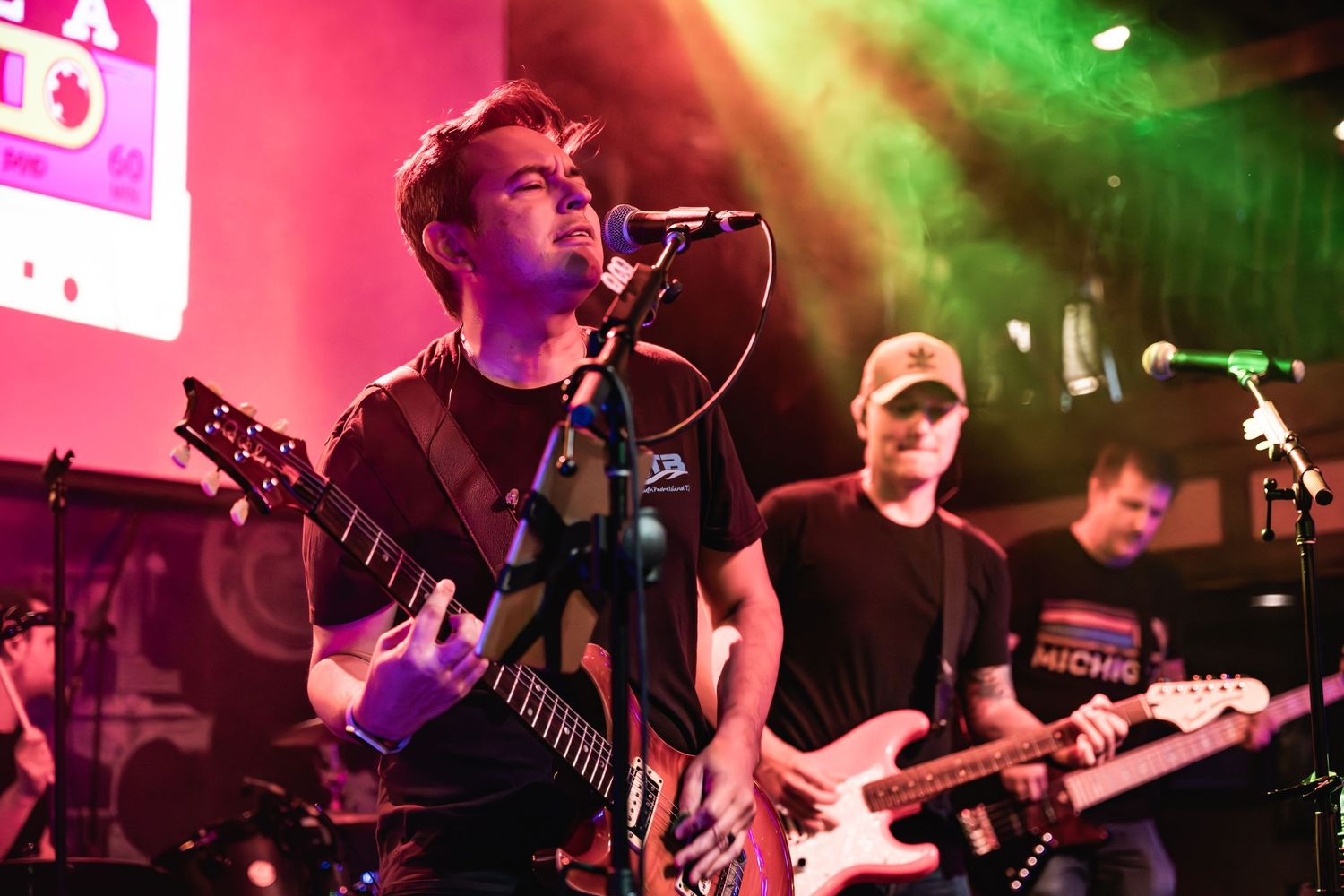 Musicians play electric guitars under vibrant pink and green stage lights during live performance.