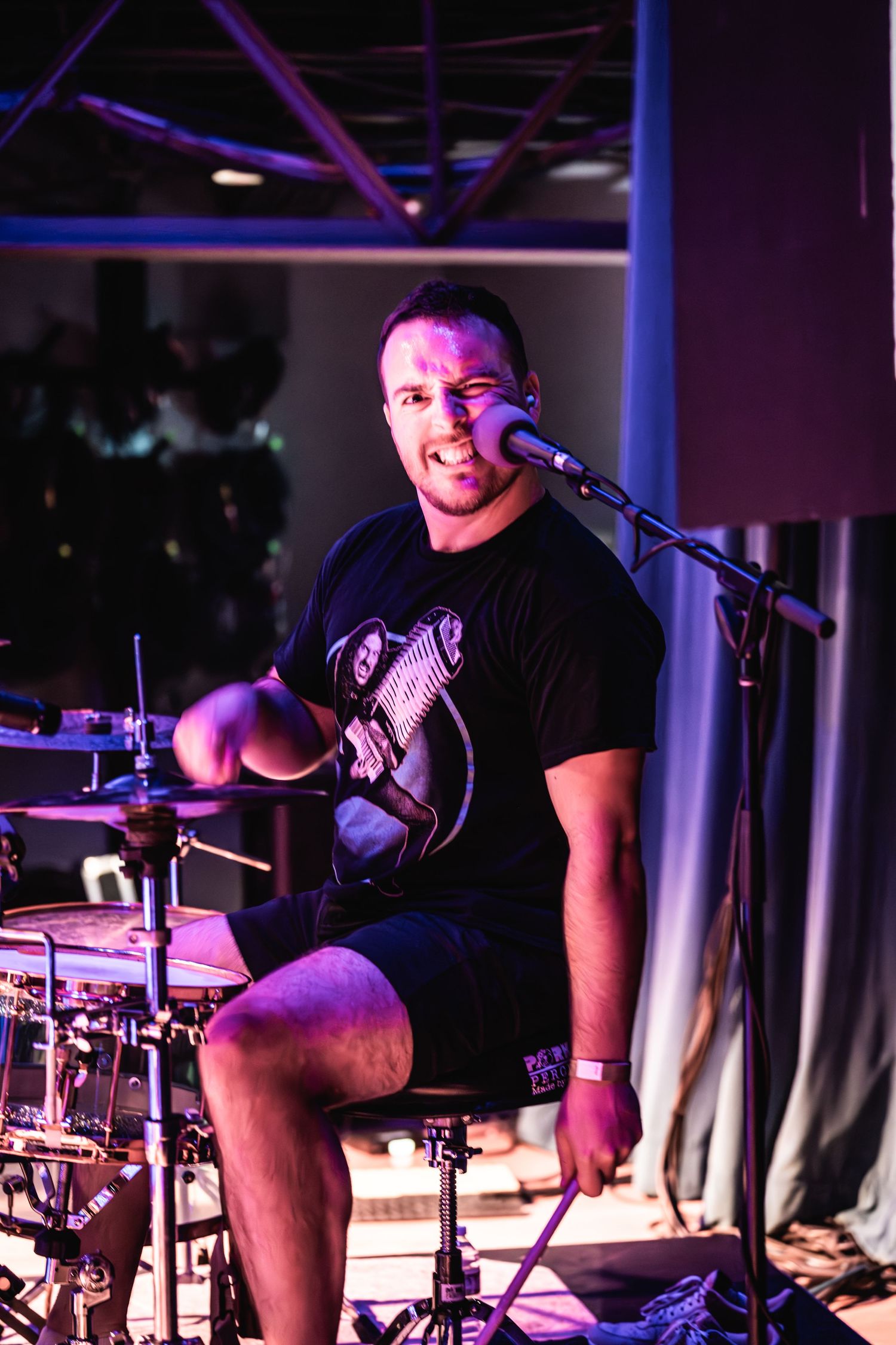 Drummer performs energetically wearing a black t-shirt under purple stage lighting.