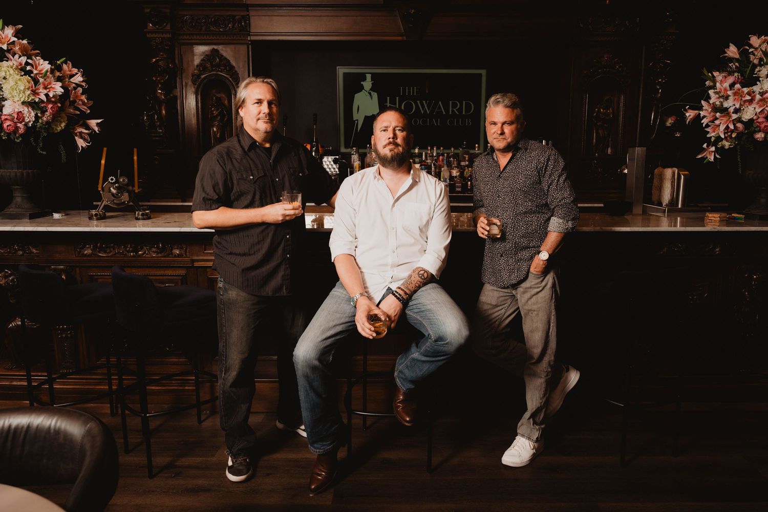 Three men posing casually at a bar counter with floral decorations.