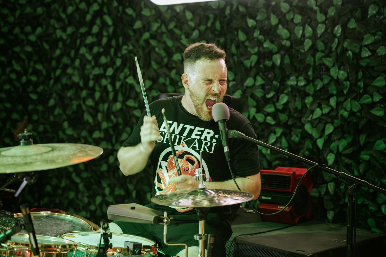 Drummer performs energetically against a green leaf backdrop during live show.