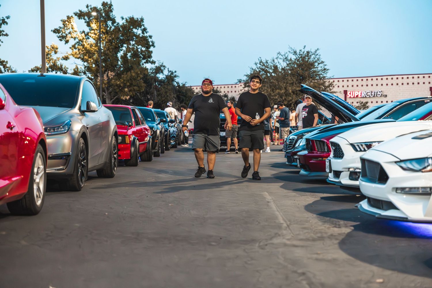 Car enthusiasts walking between rows of modified vehicles at outdoor auto meet.