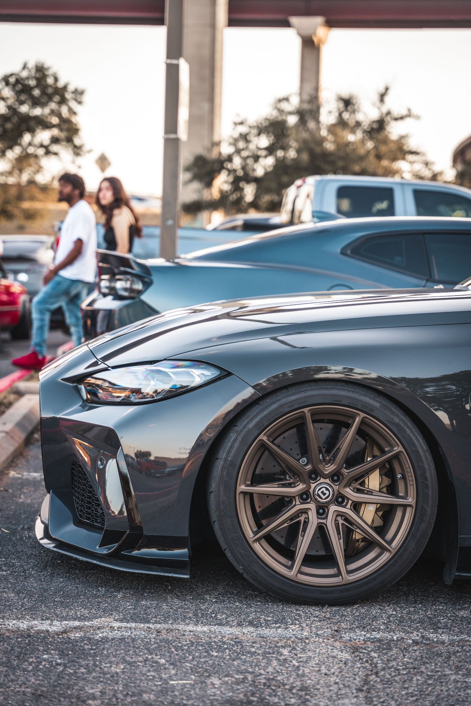 Close-up view of a grey BMW M4's front quarter panel with bronze wheels in a parking lot at sunset.