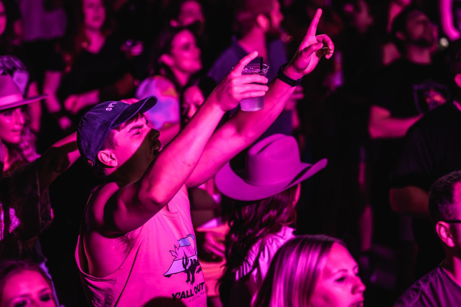 Concert crowd illuminated in pink lighting raise their hands and phones during live performance.