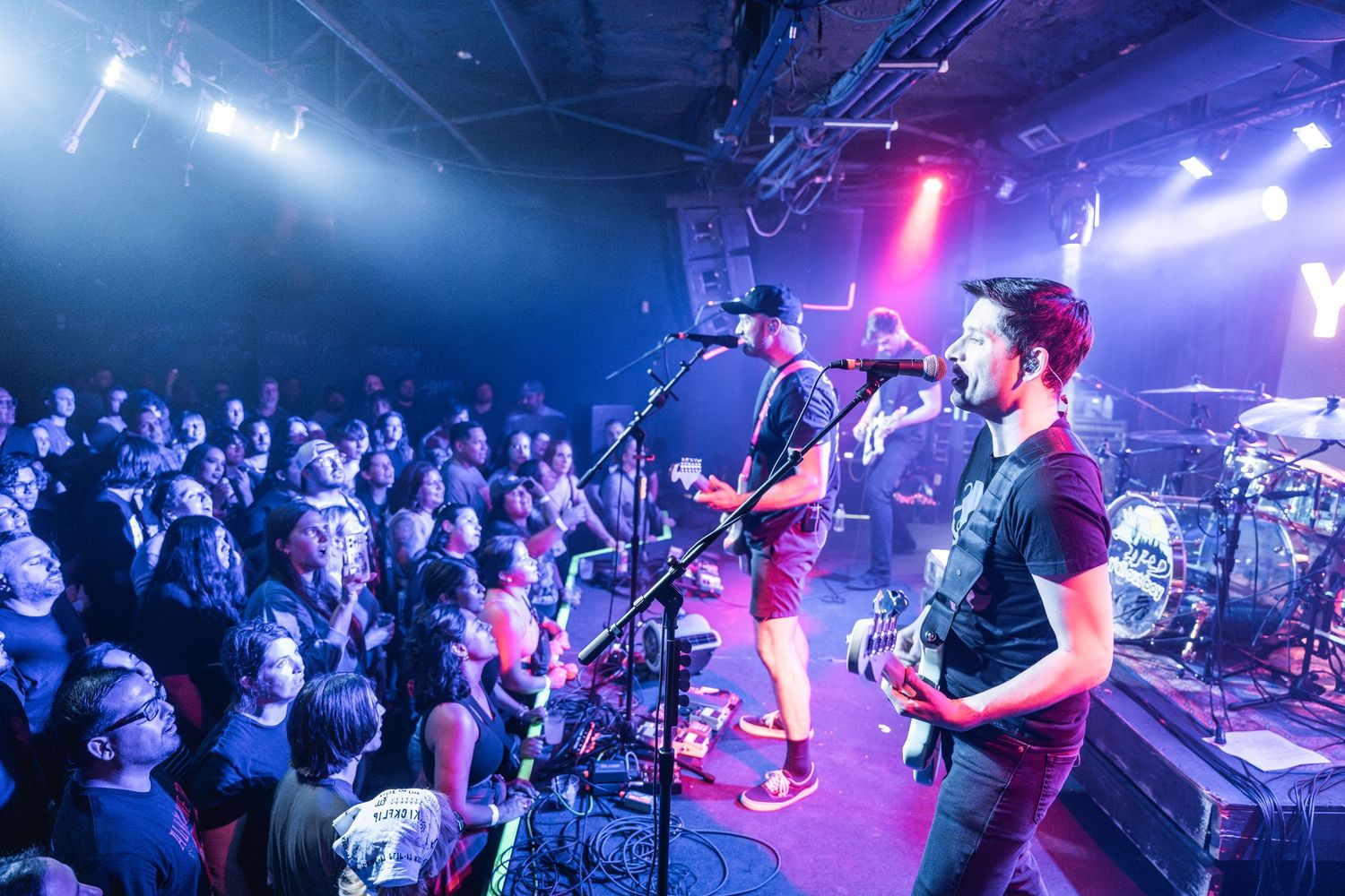 Band performs on stage with colorful blue and purple lighting while crowd watches at a packed indoor music venue.