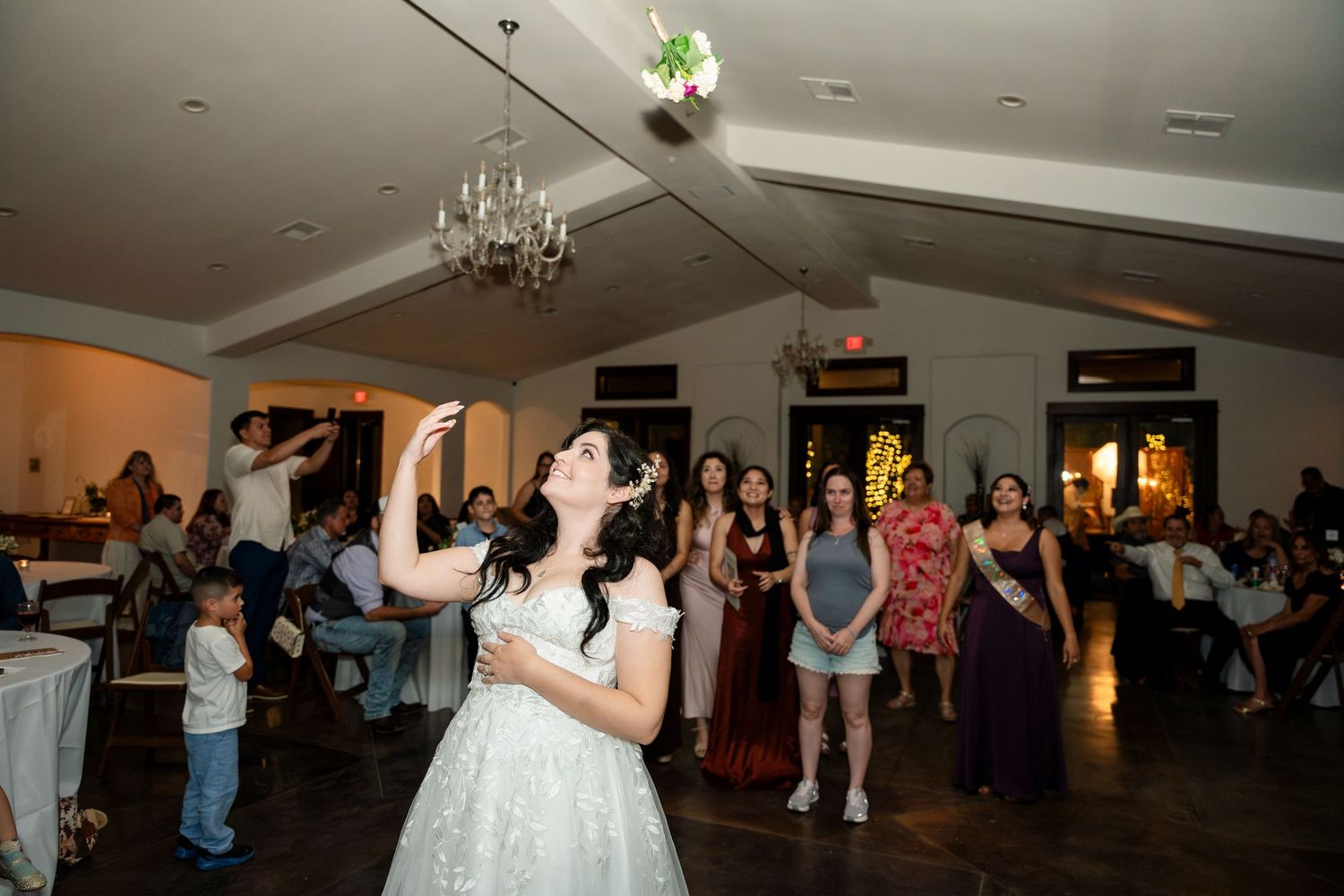 A crowd gathers on a dance floor under a chandelier as a wedding bouquet is tossed through the air.