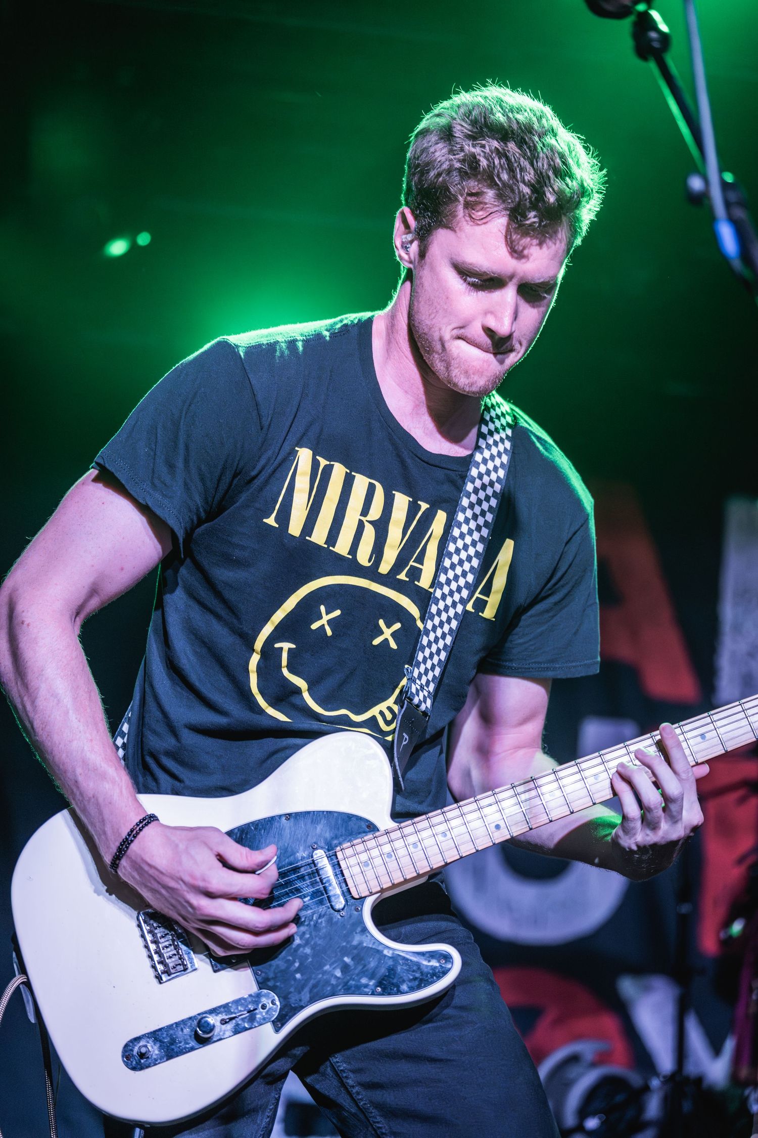 Musician in Nirvana t-shirt plays white electric guitar under green stage lighting.