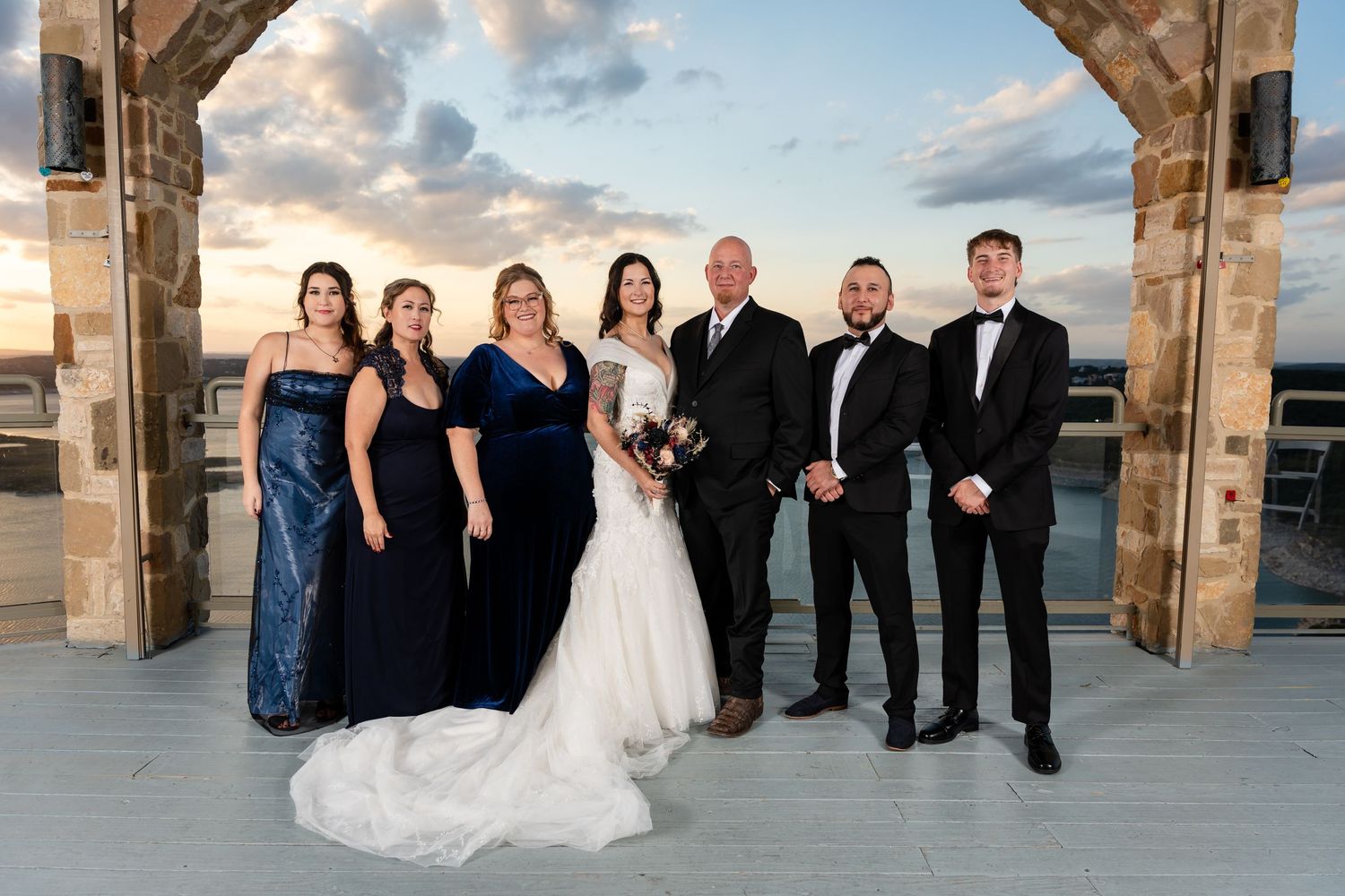 Wedding party in formal attire posed under stone archway with ocean view.