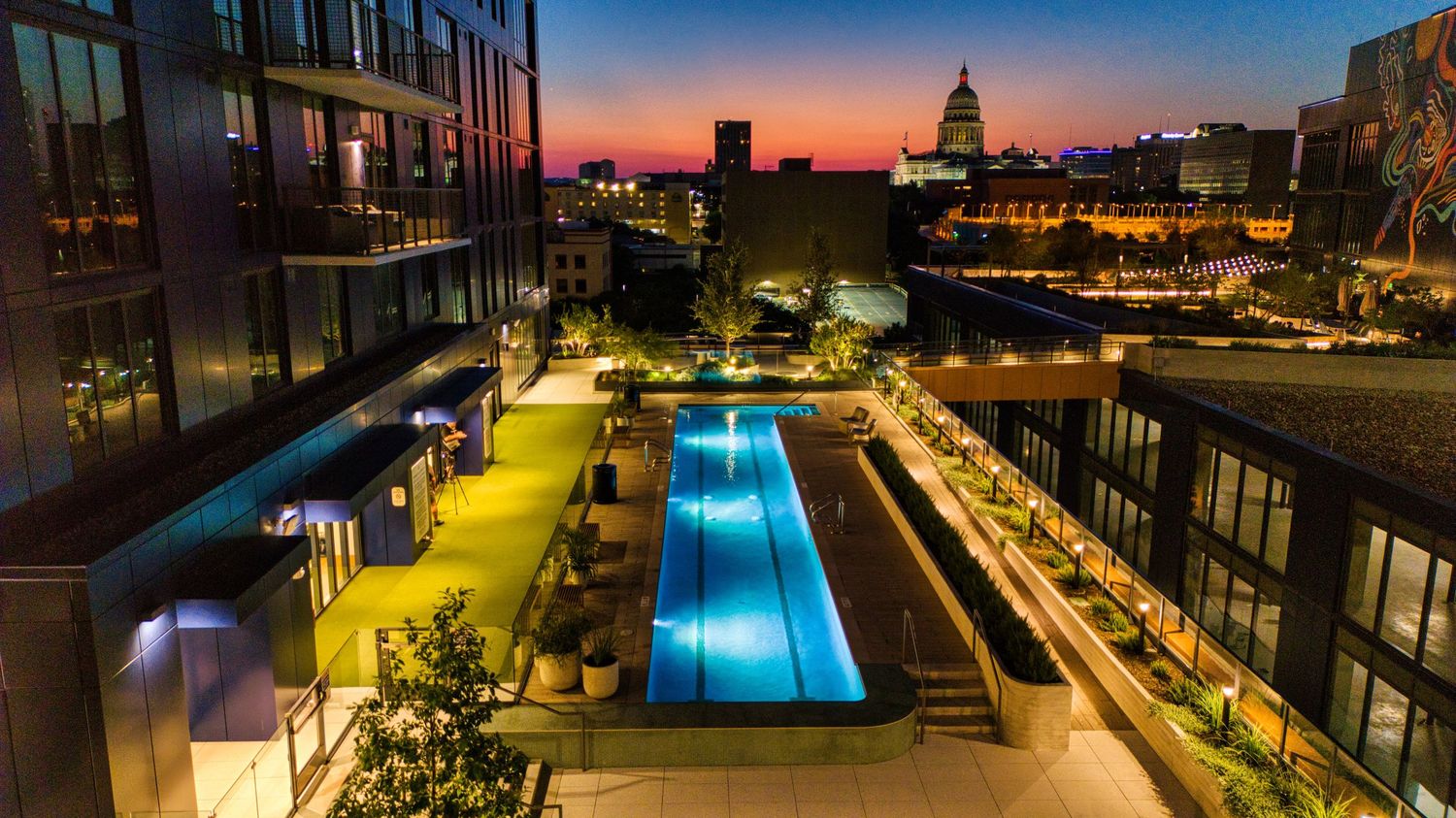 Night view of illuminated lap pool on luxury apartment building rooftop with city skyline backdrop.