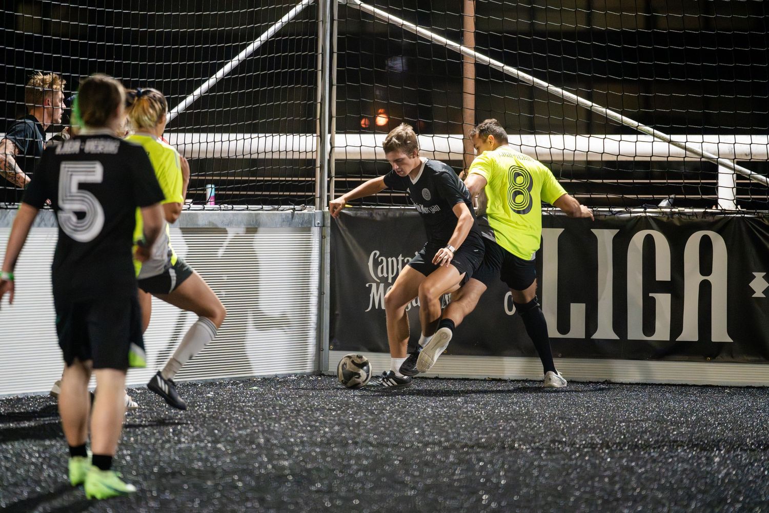 Players compete in an intense indoor soccer match with one player jumping to block a shot.