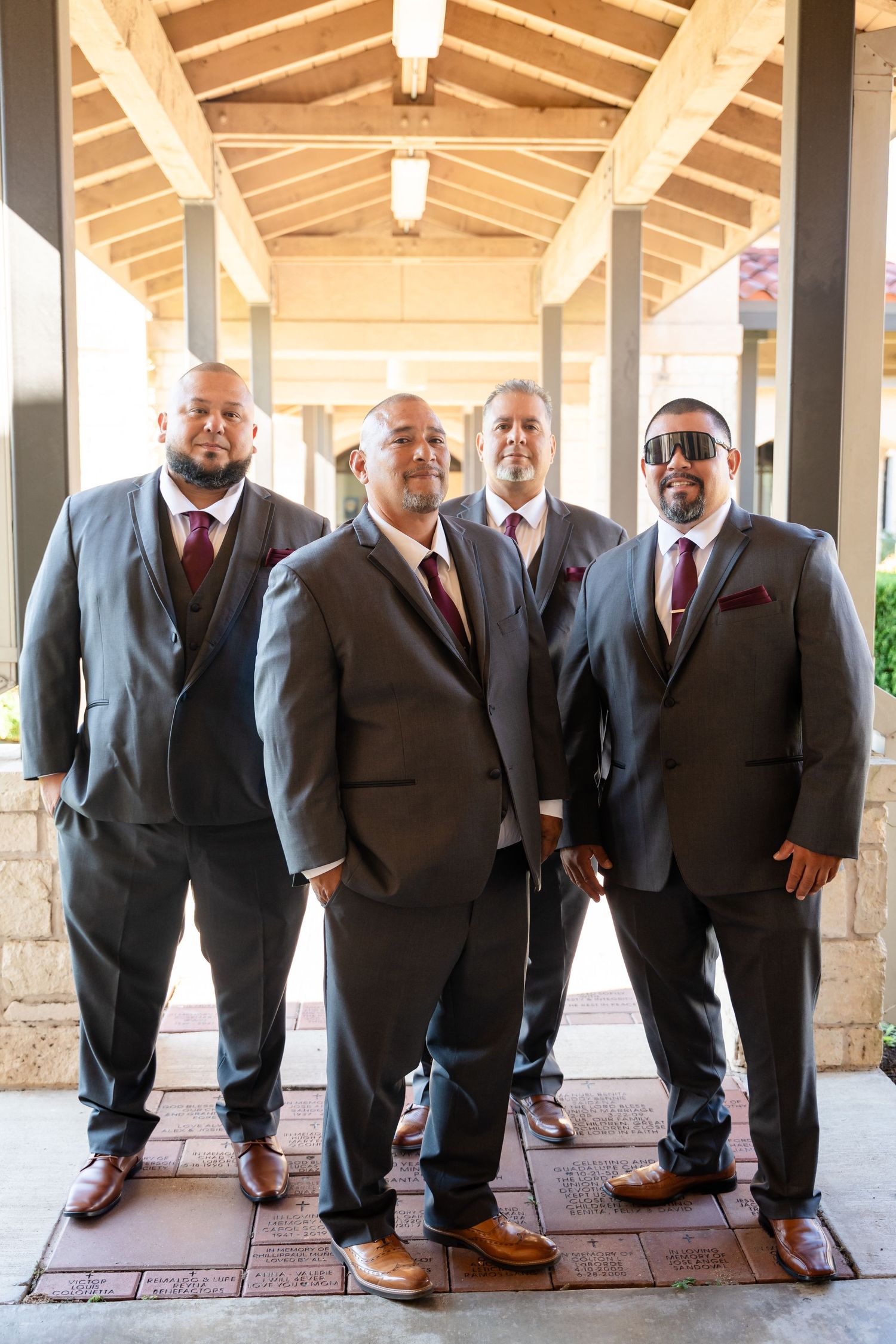 Groomsmen in matching burgundy ties and dark suits pose under a covered walkway.