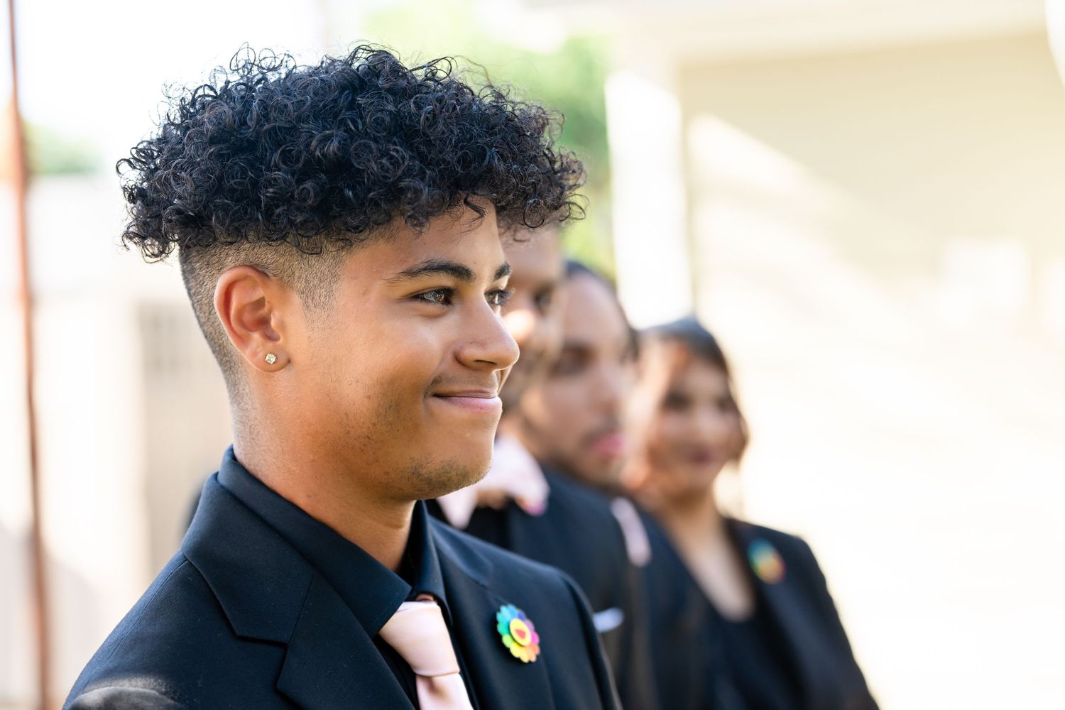 A person in formal attire with a pink tie and rainbow pin smiles during an event.