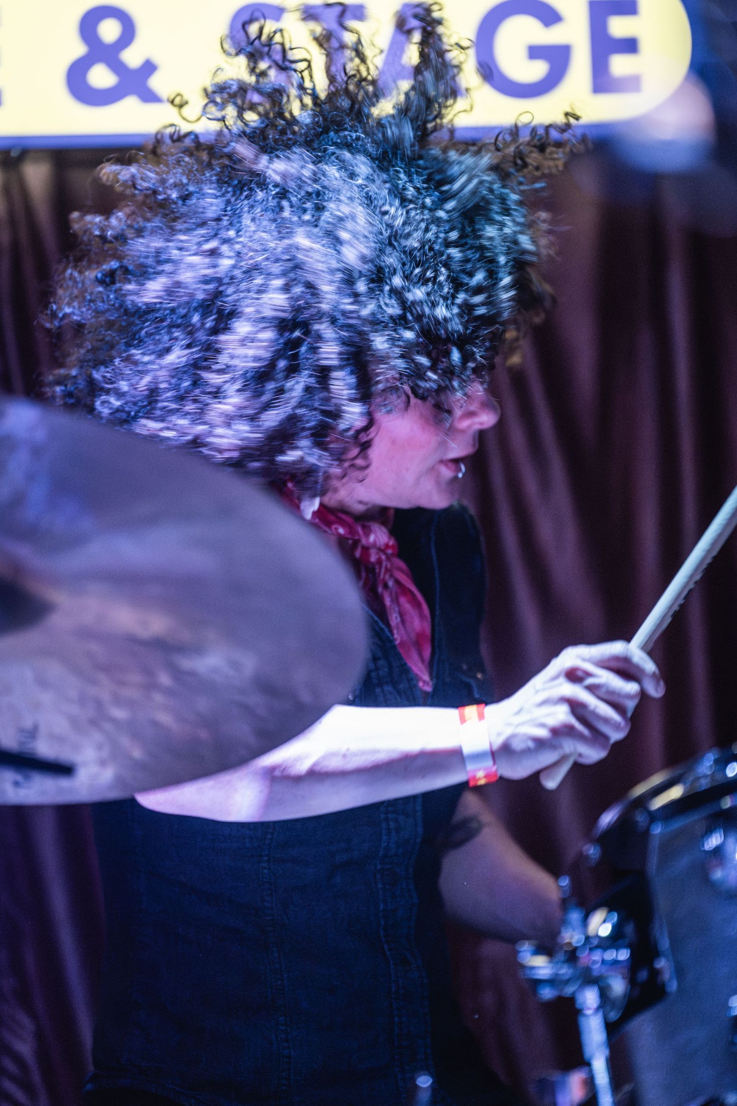A drummer with wild curly hair performs energetically on stage under blue lighting at a music venue.