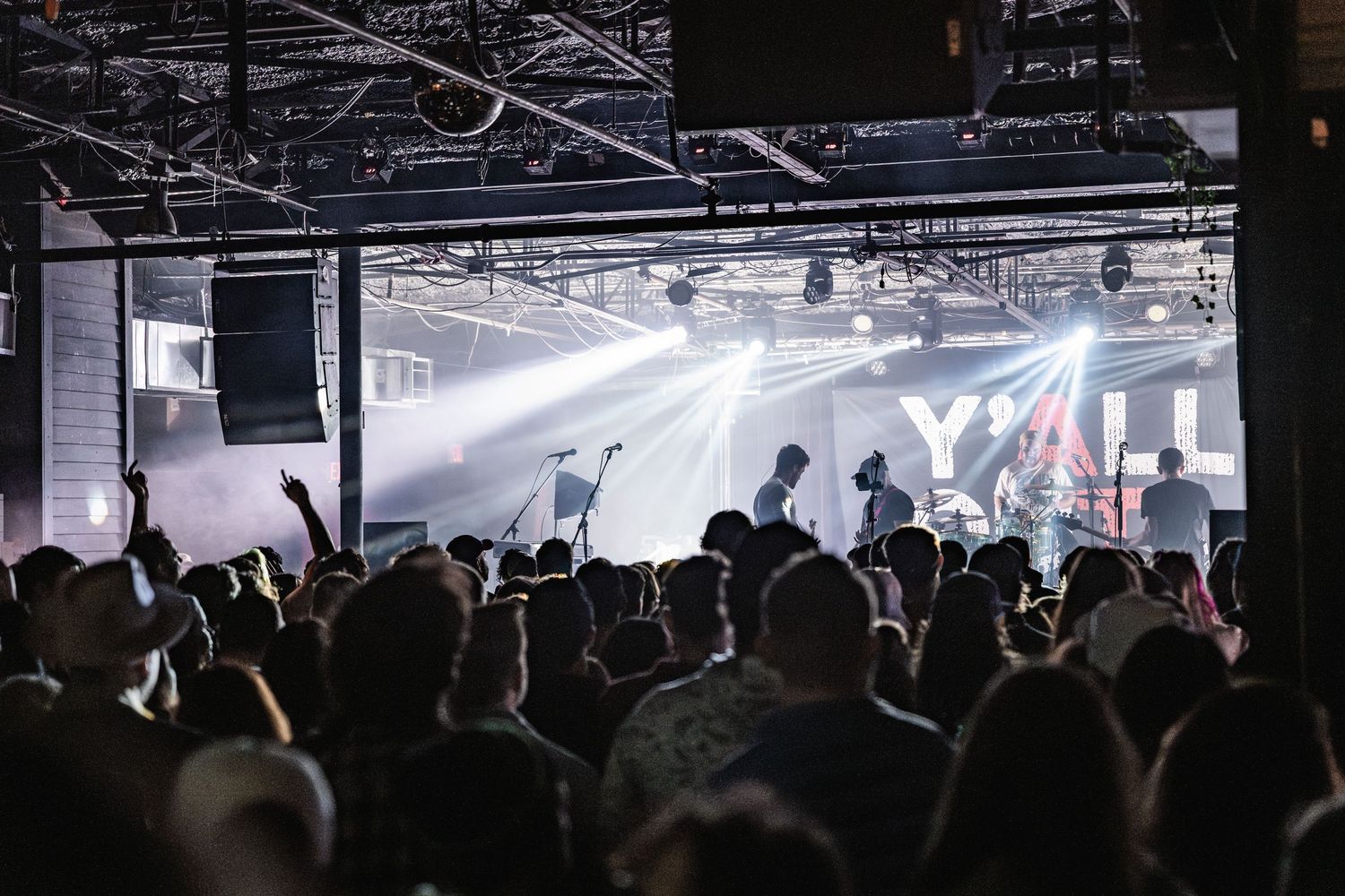 Silhouetted crowd watches a performance with bright stage lights and fog at an indoor concert venue.