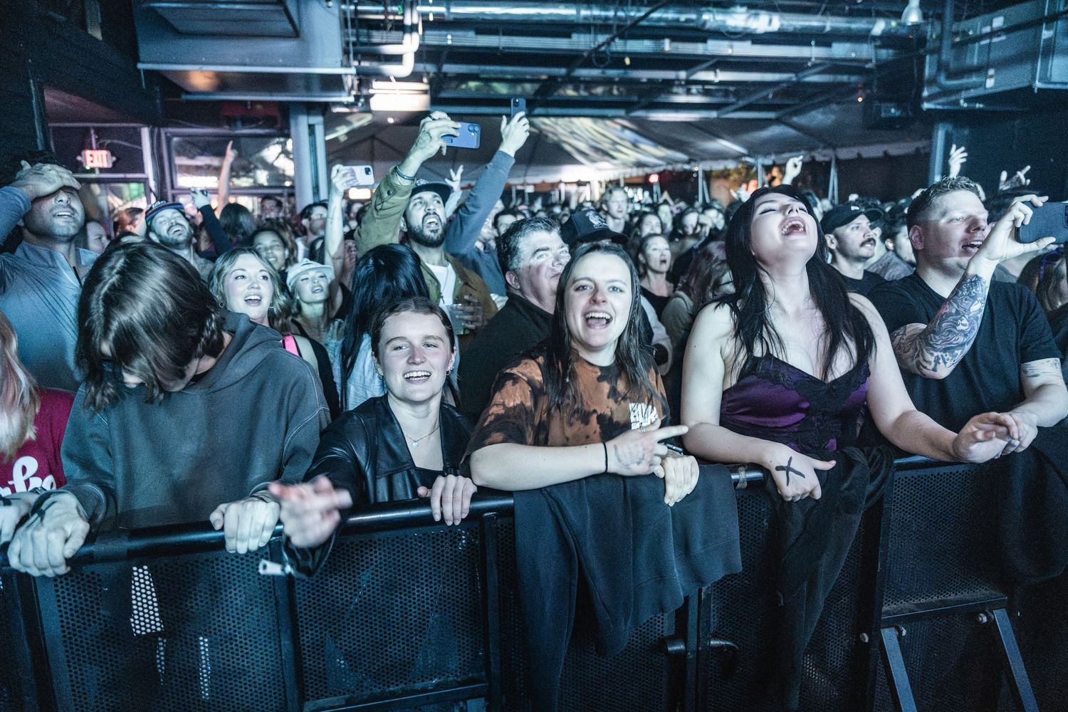 Excited concert crowd leans against metal barriers at a dark indoor venue during a live music performance.