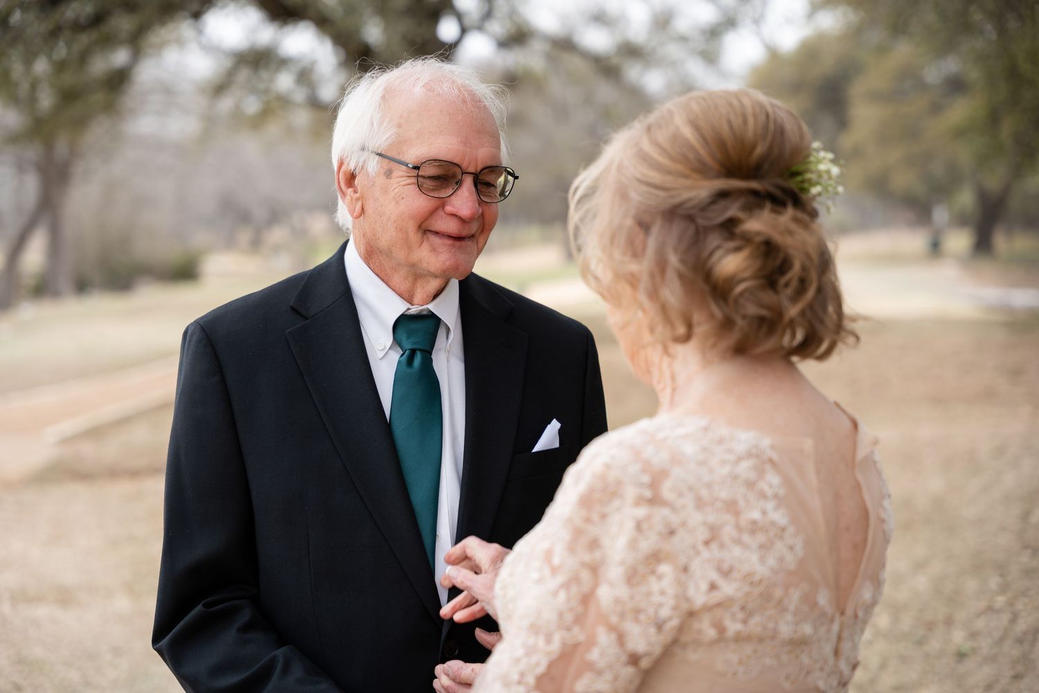 A senior man in a dark suit and teal tie sharing an emotional first look with someone in a lace dress outdoors.