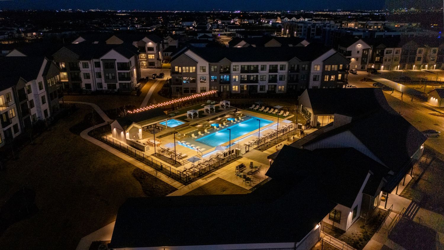 Aerial nighttime view of an illuminated apartment complex with a glowing blue swimming pool and deck area.