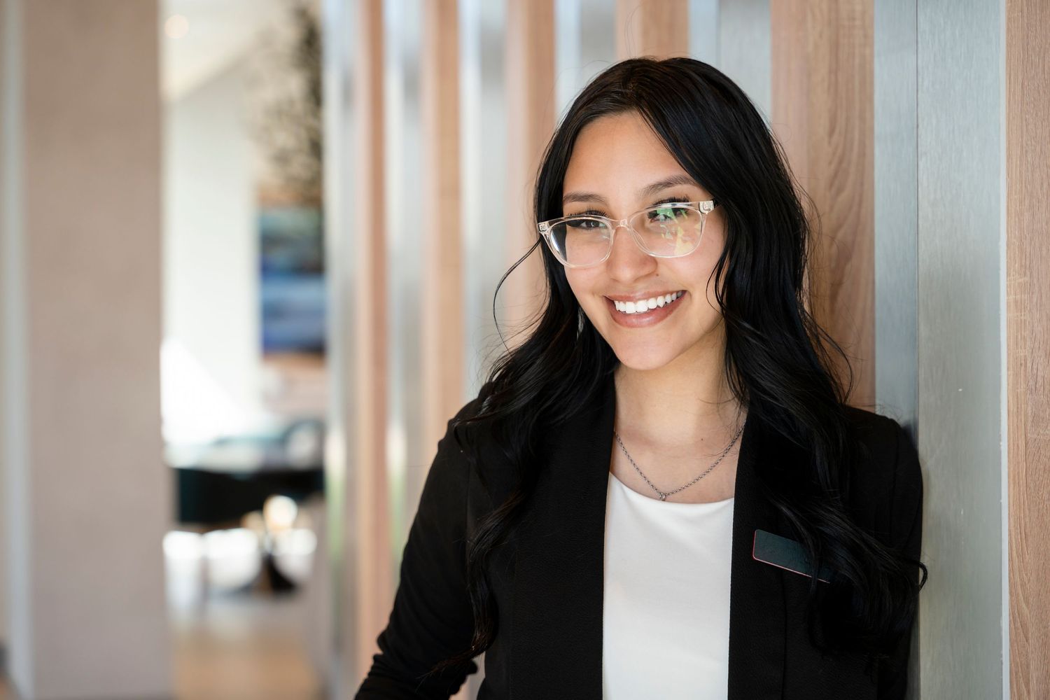 Professional wearing glasses and black blazer stands confidently in office hallway with warm lighting.