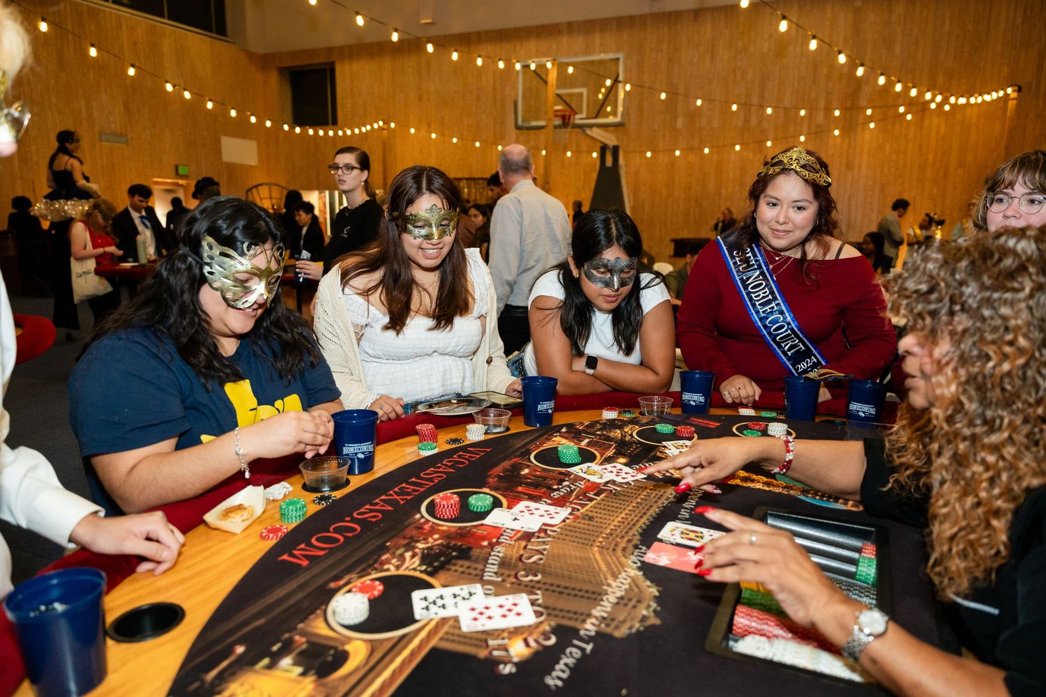 Group of people playing casino games at a red gaming table under festive string lights.