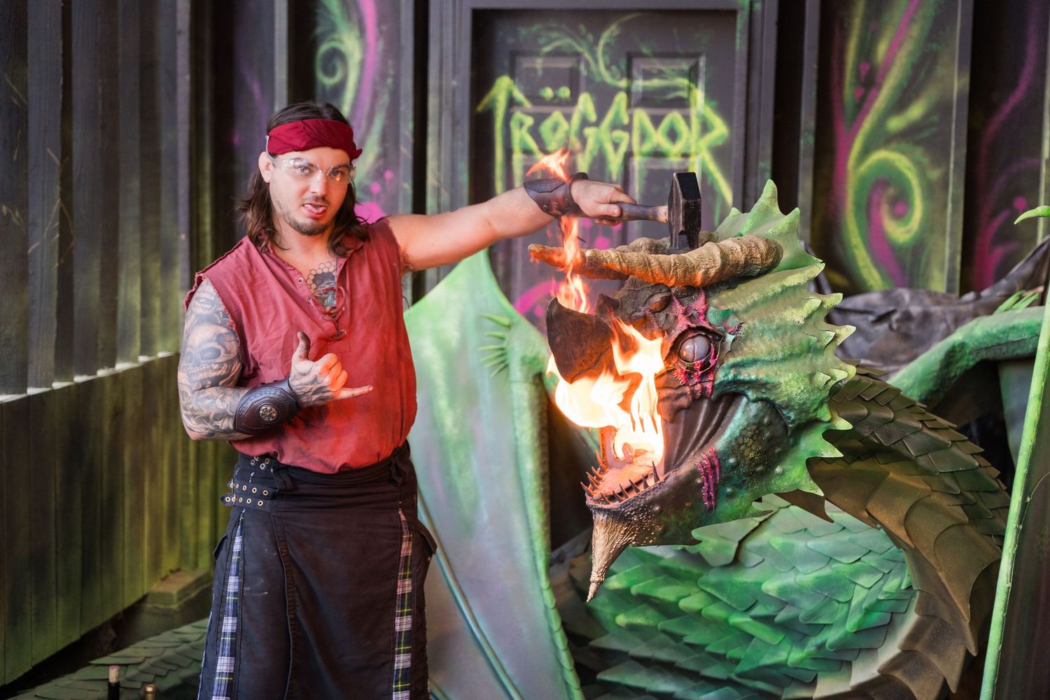 Fire performer with red bandana manipulates flames next to a large green dragon prop during a medieval show.