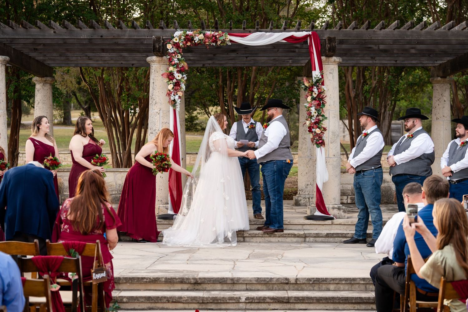 A wedding ceremony under a decorated pergola with burgundy and white floral arrangements and draped fabric.
