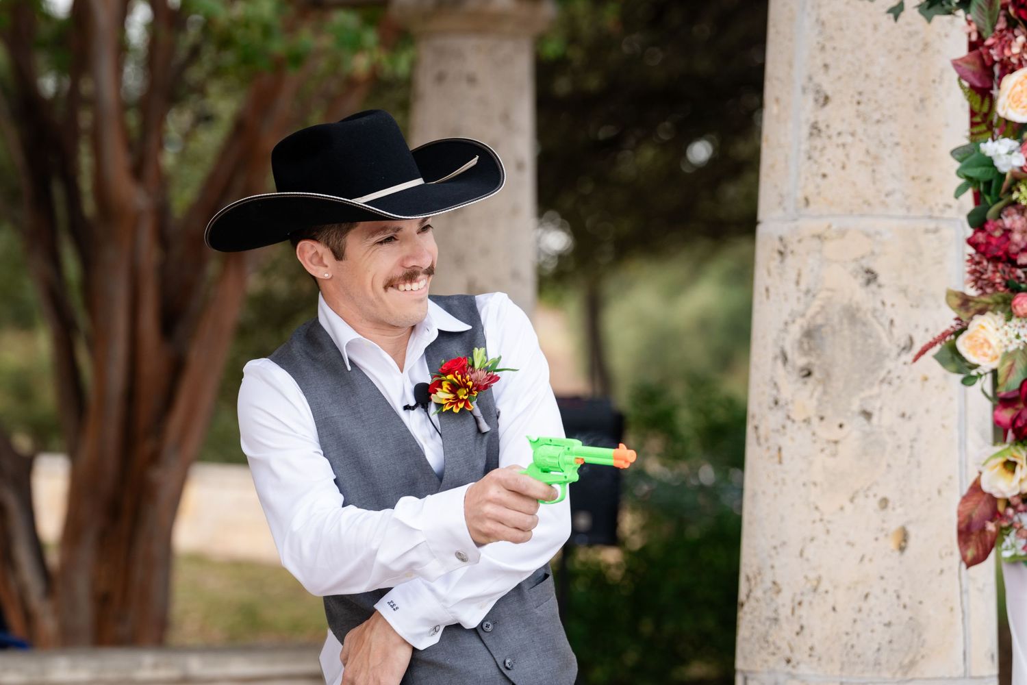 Person in western wedding attire smiles while holding a colorful drink during outdoor celebration.