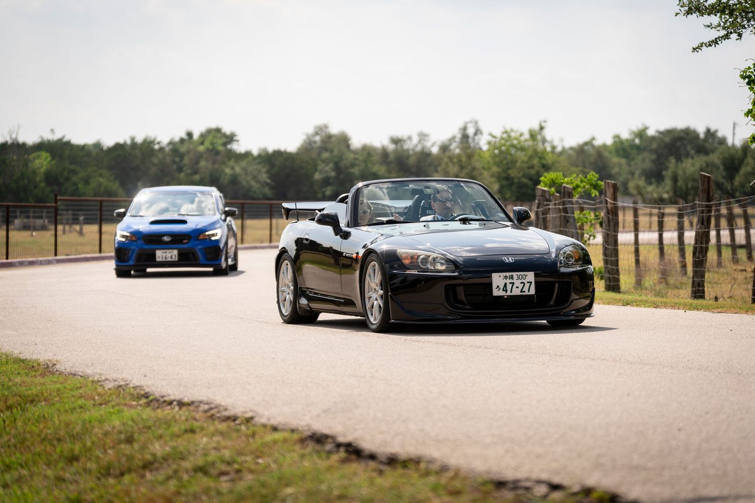 Two Japanese sports cars, a black Honda S2000 and blue Subaru WRX, driving on a winding country road.
